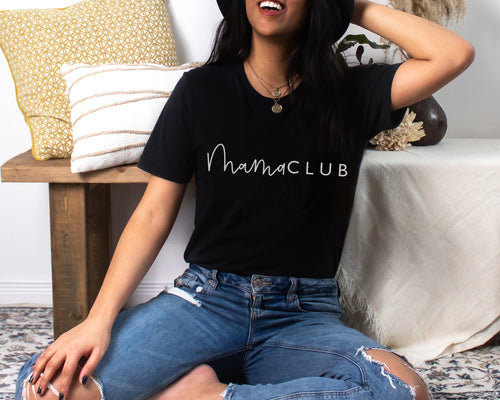 Female model smiling, sitting on a patterned rug infront of a wooden bench with cushions on. The model is wearing ripped jeans and black t-shirt with a white 'mama club' logo in a mixture of script and plain font