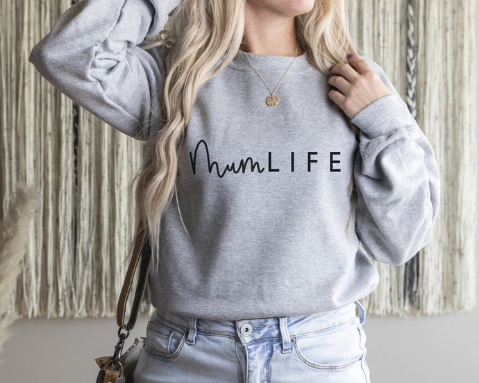 Female model wearing a grey sweatshirt with rolled up sleeves featuring 'mum life' black logo in a mixture of script and font. The background is off white with a string art wall hanging and pampas grass.
