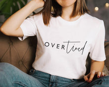 Load image into Gallery viewer, Female model with short brown hair sitting on a brown chair wearing blue jeans and a white t-shirt featuring a logo which reads &#39;Over Tired&#39; in a mixture of script and plain font 
