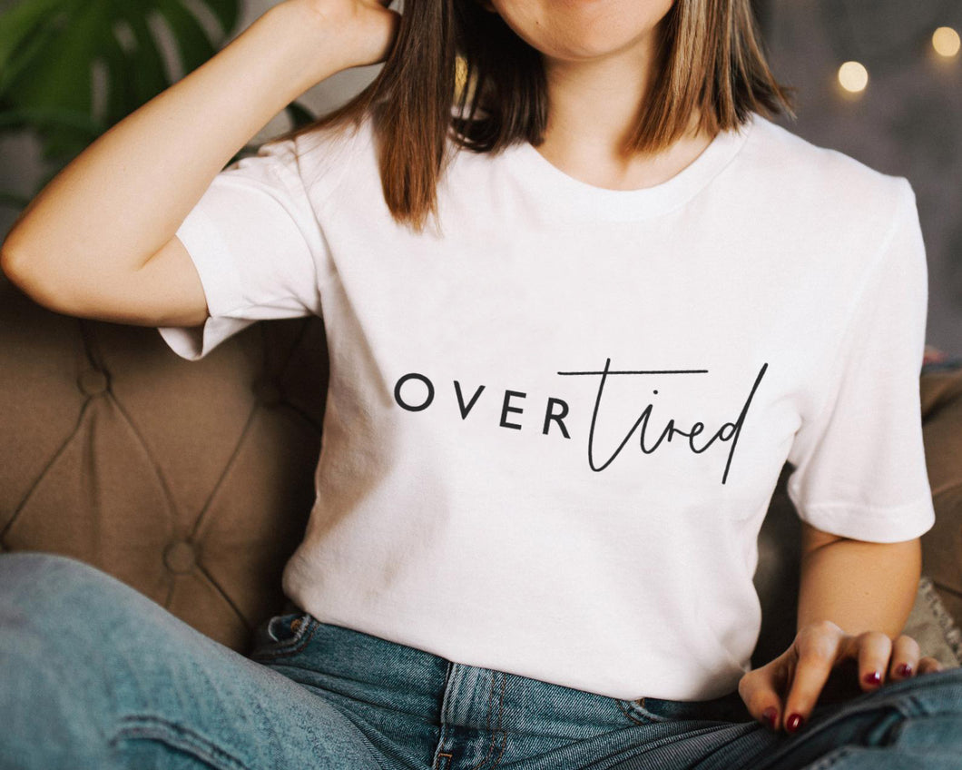 Female model with short brown hair sitting on a brown chair wearing blue jeans and a white t-shirt featuring a logo which reads 'Over Tired' in a mixture of script and plain font 